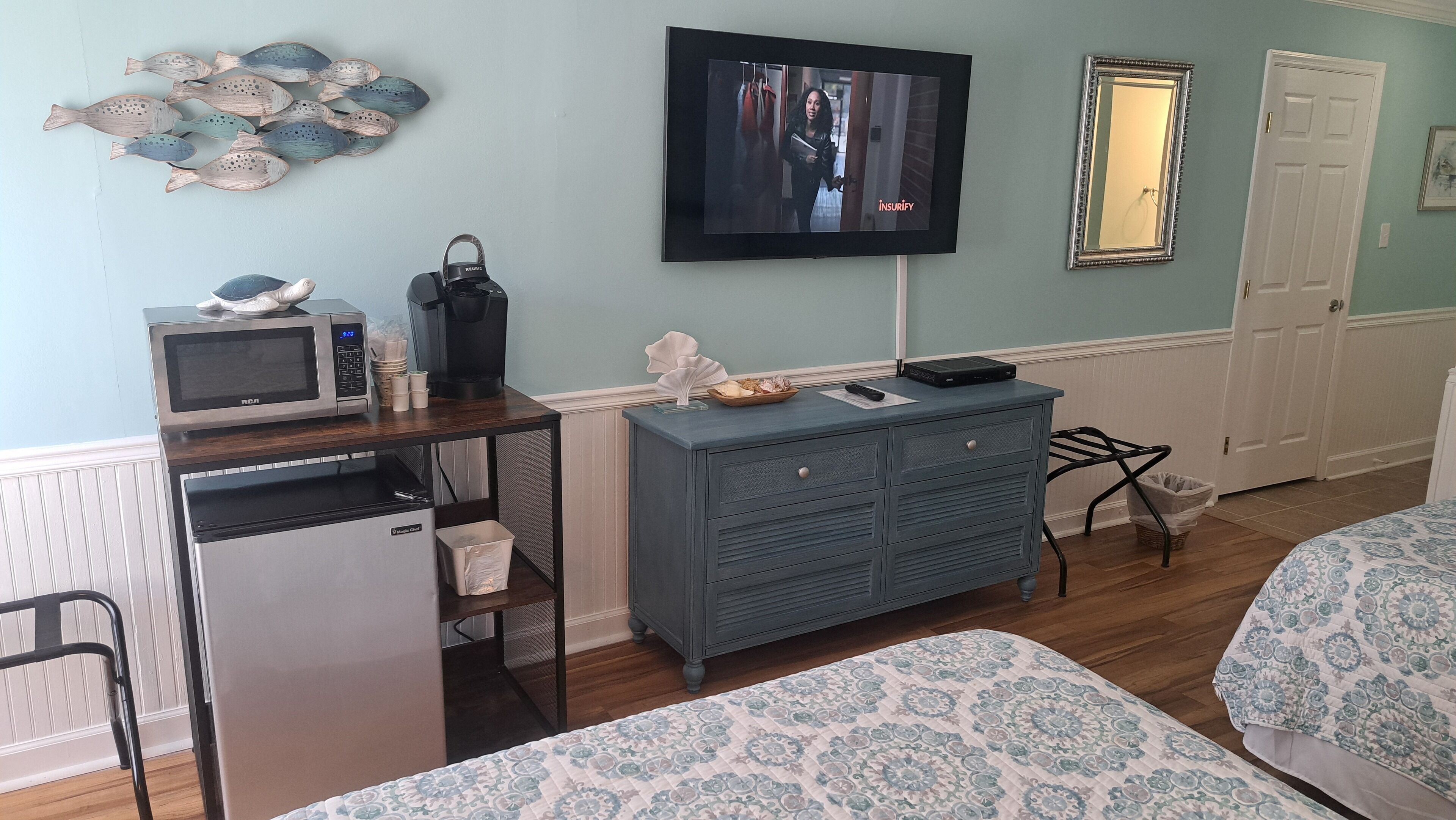 A compact hotel room with a kitchenette, a flat-screen TV, a dresser, and two beds with patterned bedding.
