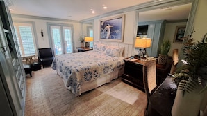 A bedroom with a large bed, a nightstand, a chair, and a desk with a lamp. There is a window with blinds, a door, and a painting on the wall. - Saint Simons Inn by the Lighthouse (St. Simons Island)