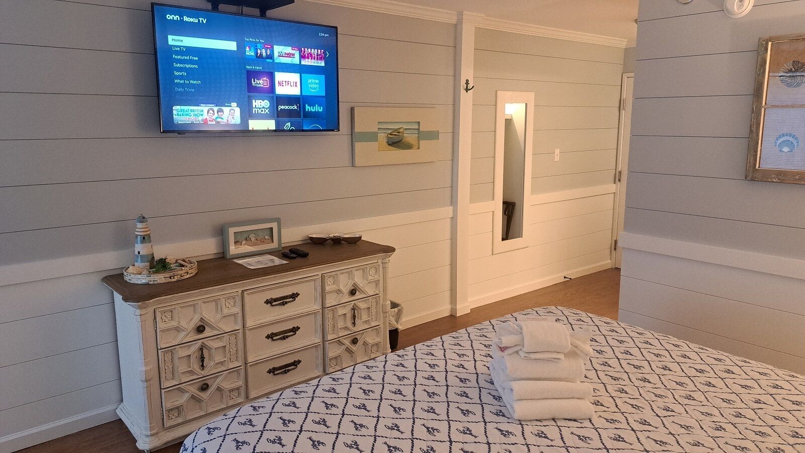 Habitación de hotel con televisión montada en la pared, cómoda con cajones, cama con colcha estampada y espejo con marco decorativo.