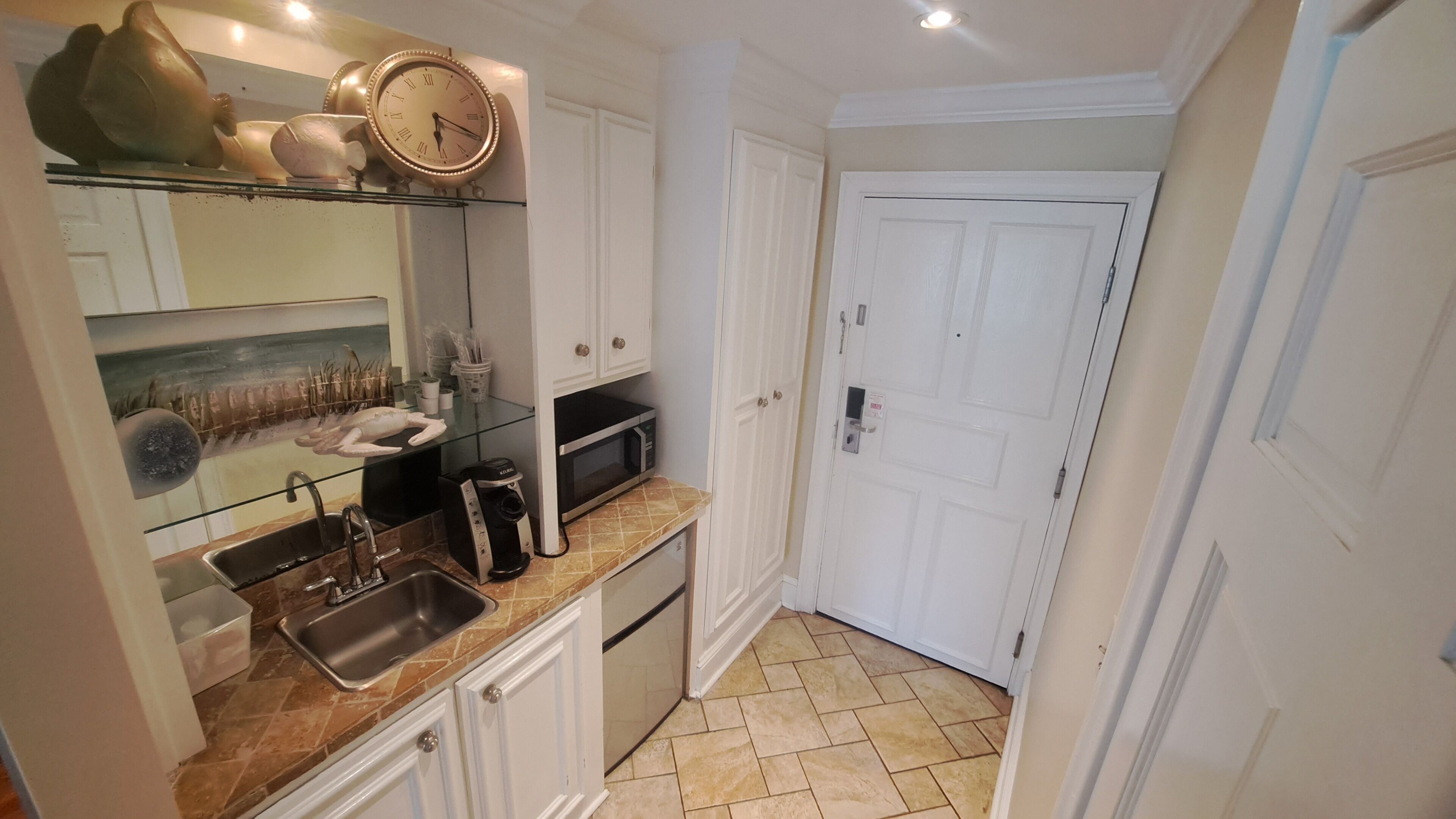 Cocina compacta con un lavamanos, microondas y cafetera. Se observa una puerta con manija y un armario blanco con repisa de vidrio arriba.