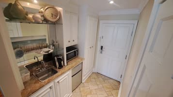 Cocina compacta con un lavamanos, microondas y cafetera. Se observa una puerta con manija y un armario blanco con repisa de vidrio arriba.