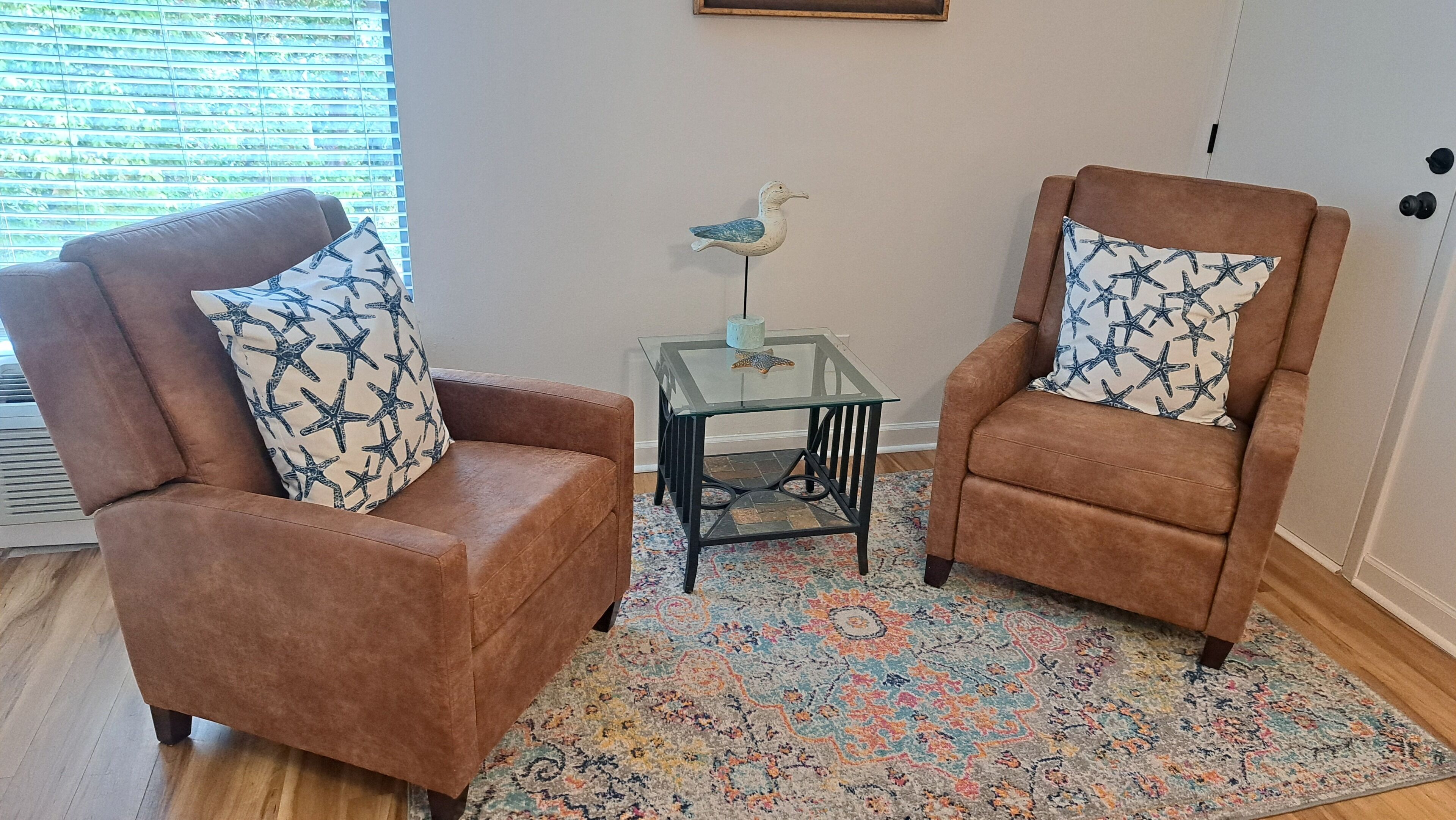 Deux fauteuils marron ornés de coussins à motifs d’étoiles de mer, une table en verre sur laquelle est posée une statuette d’oiseau décorative, et un tapis aux couleurs vives.