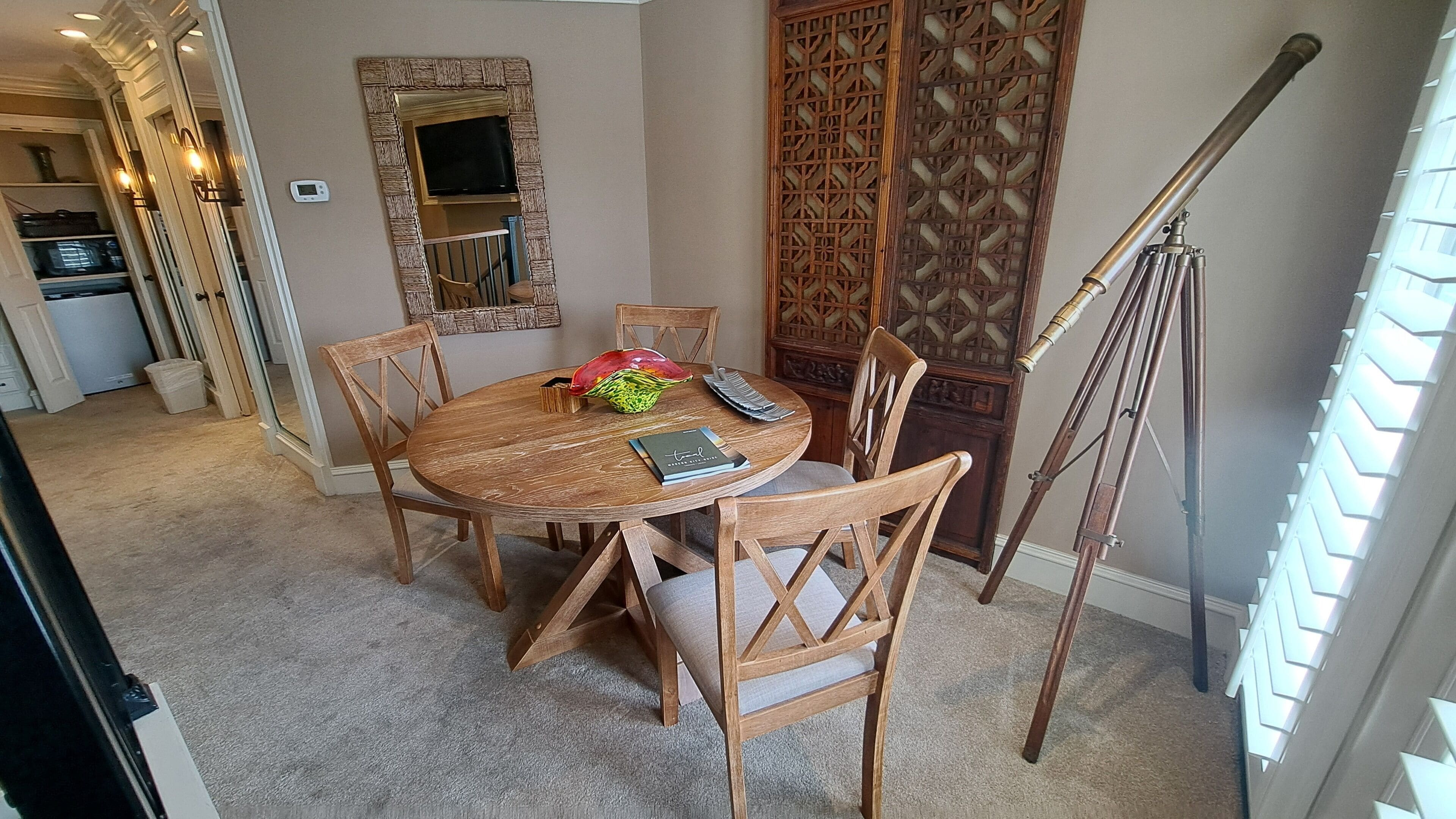 A dining area with a round wooden table, four chairs, a decorative mirror, a telescope, and a patterned screen.