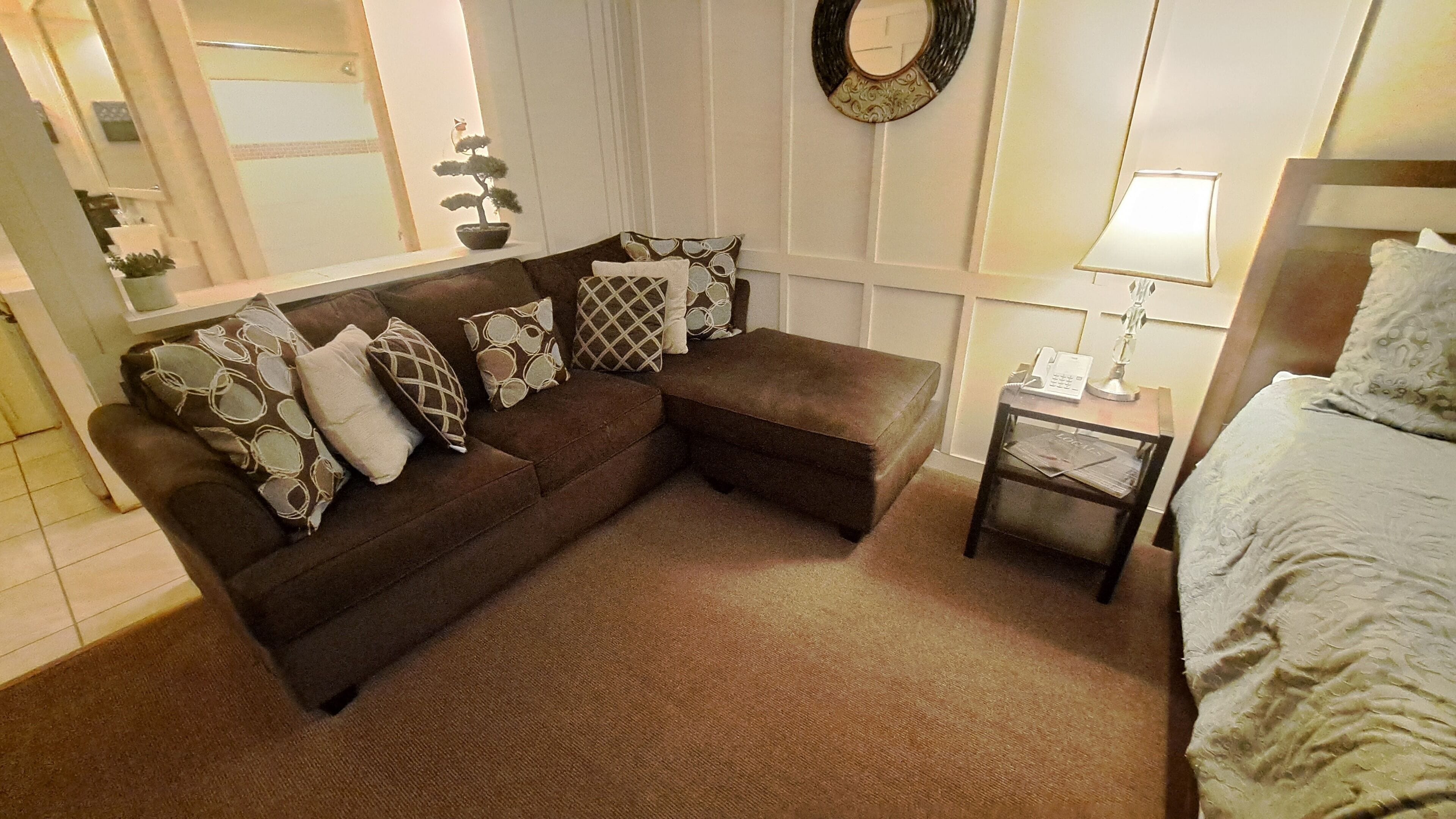 A cozy living room with a brown sectional sofa, patterned cushions, a small side table with a lamp, and a bed with a white comforter.