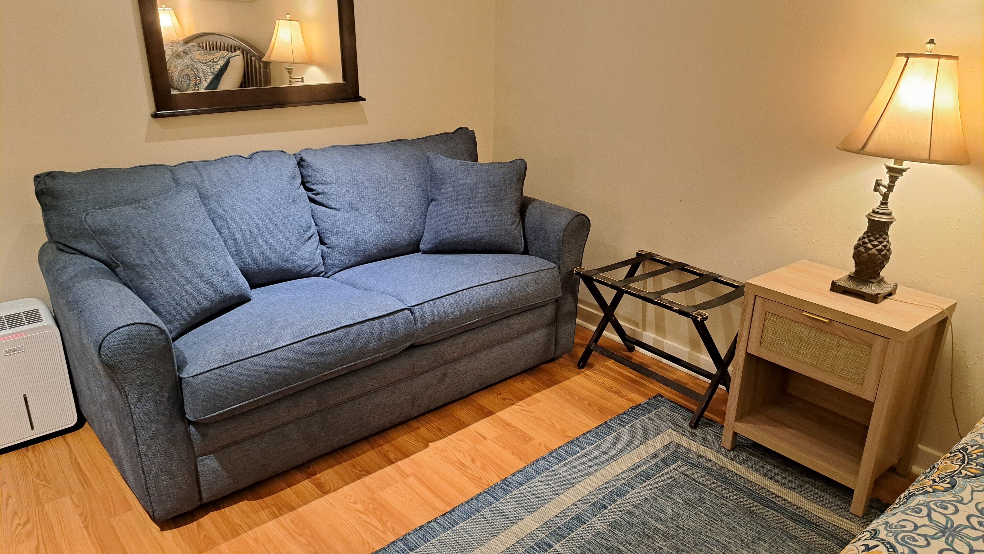 Una habitación con un sofá gris, una pequeña mesa de noche de madera con una lámpara, un espejo y un tapete estampado.