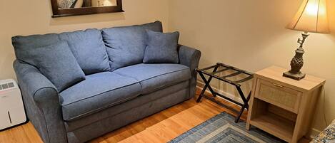 A room with a grey sofa, a small wooden side table with a lamp, a mirror, and a patterned rug.