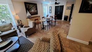 A living room with a sofa, armchairs, a coffee table, and a dining area with chairs and a table. There is a painting on the wall, a ceiling fan, and a window with a view of trees. - Saint Simons Inn by the Lighthouse (St. Simons Island)
