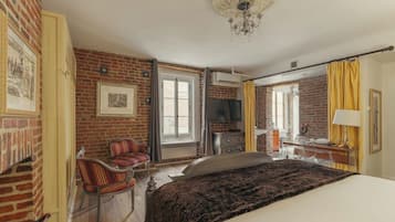 A hotel room with a brick wall, a bed with a dark blanket, a TV, a desk with a lamp, and a chair.