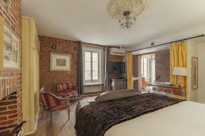 A hotel room with a brick wall, a bed with a dark blanket, a TV, a desk with a lamp, and a chair.