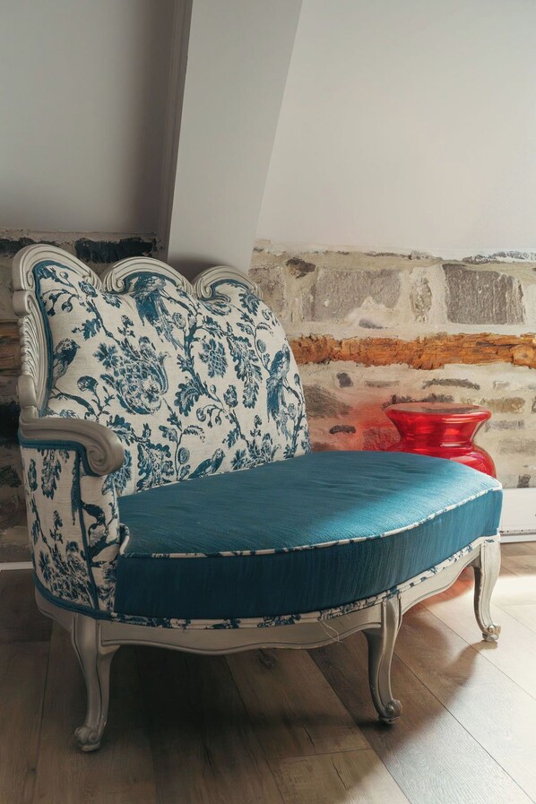 Un canapé de style vintage, orné d’un motif floral et agrémenté d’un coussin turquoise, est placé dans une pièce dont le mur est en briques apparentes et le sol en bois.