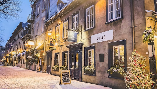 Eine schneebedeckte Kopfsteinpflasterstraße gesäumt von historischen Gebäuden, darunter ein Restaurant namens "Jules" und ein Schild für "Le Place d'Armes".