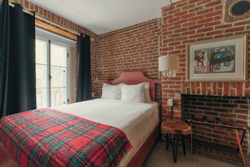 A hotel room with a brick wall, a bed with white bedding, a small table, and a fireplace.