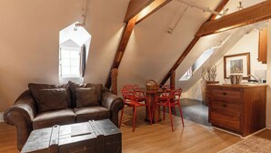 Ein Loft-Zimmer mit schrägen Wänden, einem braunen Ledersofa, einer Holzkisten-Kommode, einem Esstisch mit roten Stühlen und einem gemusterten Teppich.