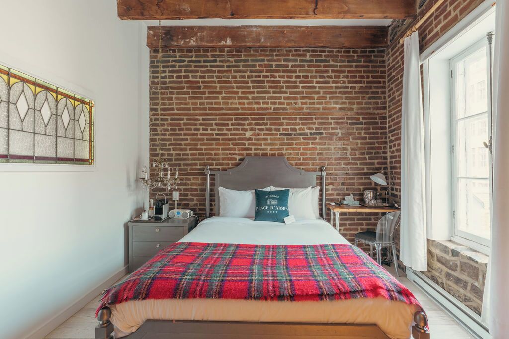 A bedroom with a brick wall, a bed with a plaid blanket, a nightstand, a desk, and a window.