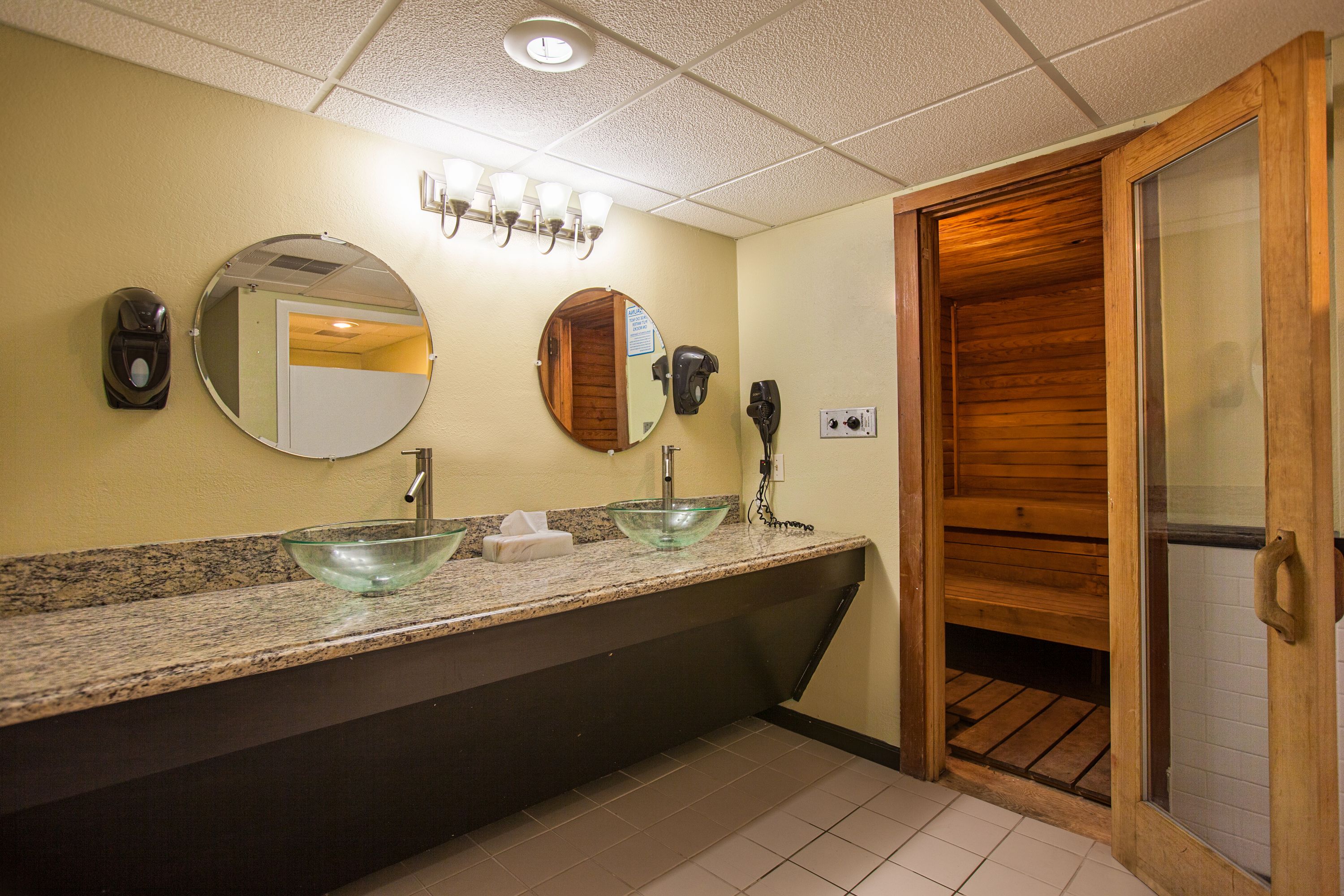 A bathroom with a granite countertop, two sinks, a round mirror, a towel dispenser, and a wooden door.