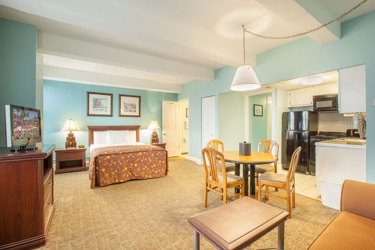A hotel room with a bed, a desk, a chair, a dining table with chairs, a kitchen area, and a television.
