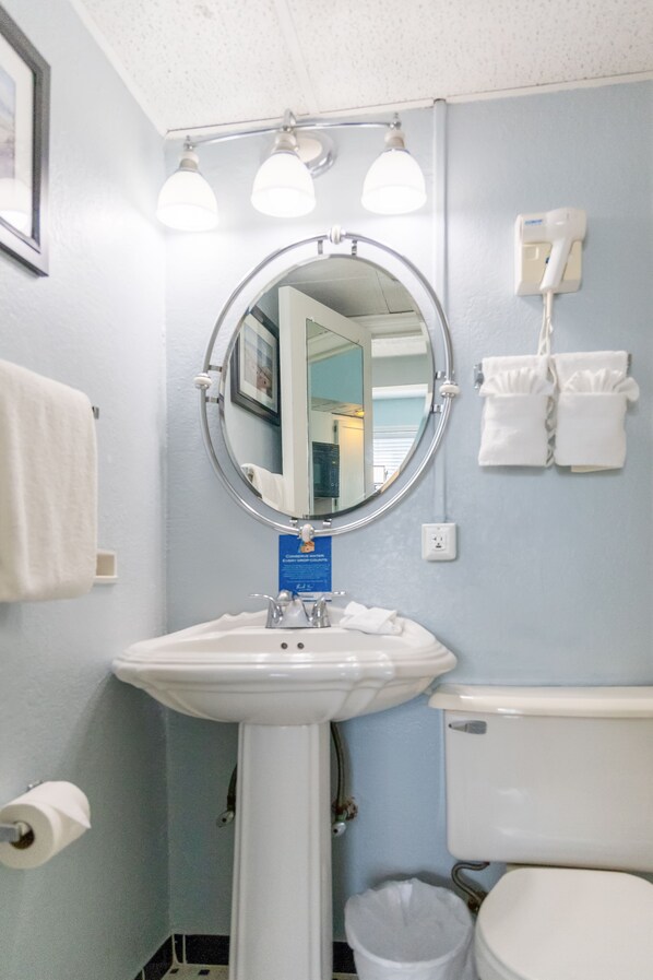 A bathroom with a sink, mirror, and toilet. There are towels hanging on the wall and a trash can.