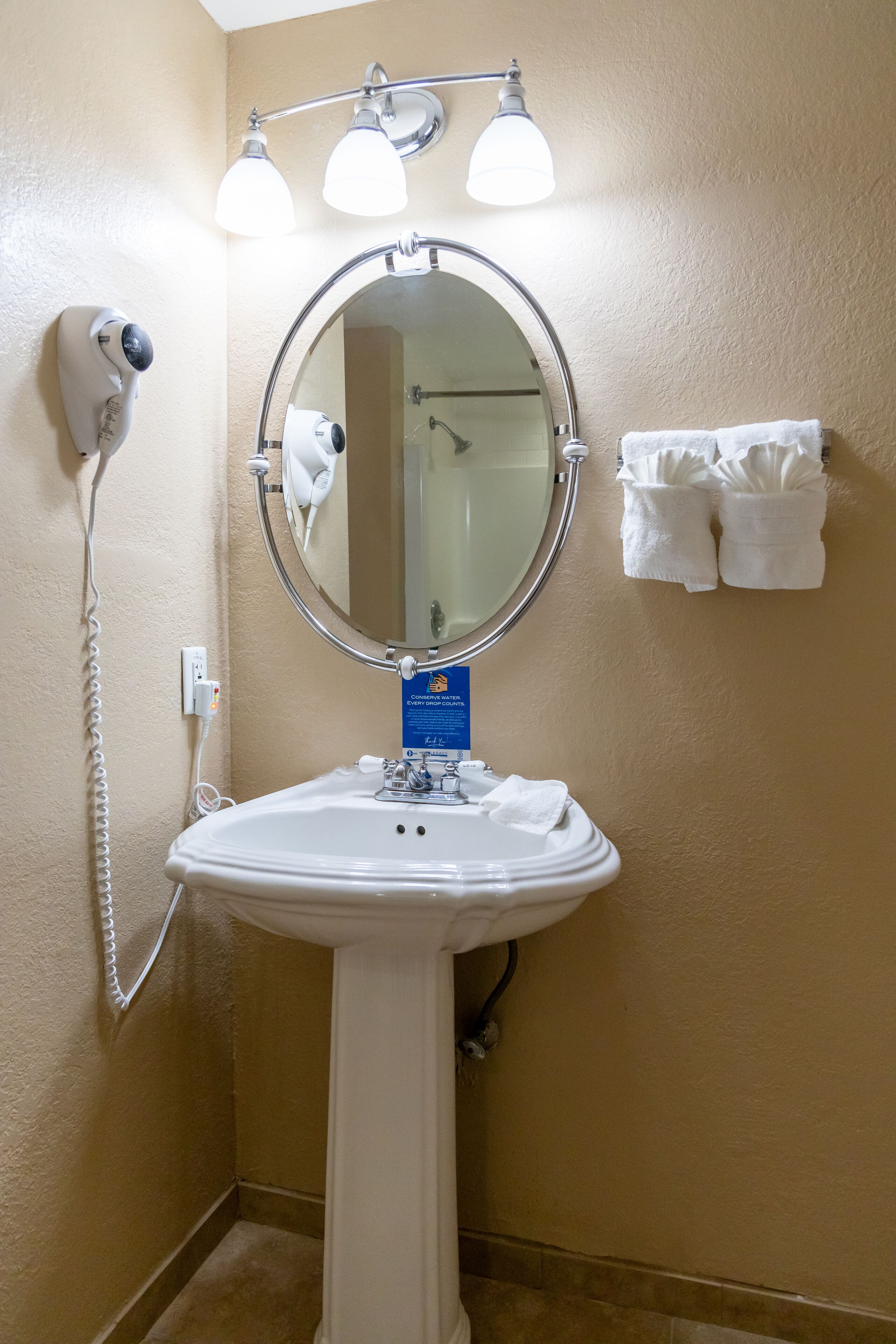 A bathroom with a white sink, a round mirror, and a hairdryer on the wall. There are towels hanging on the wall and a sign above the sink.