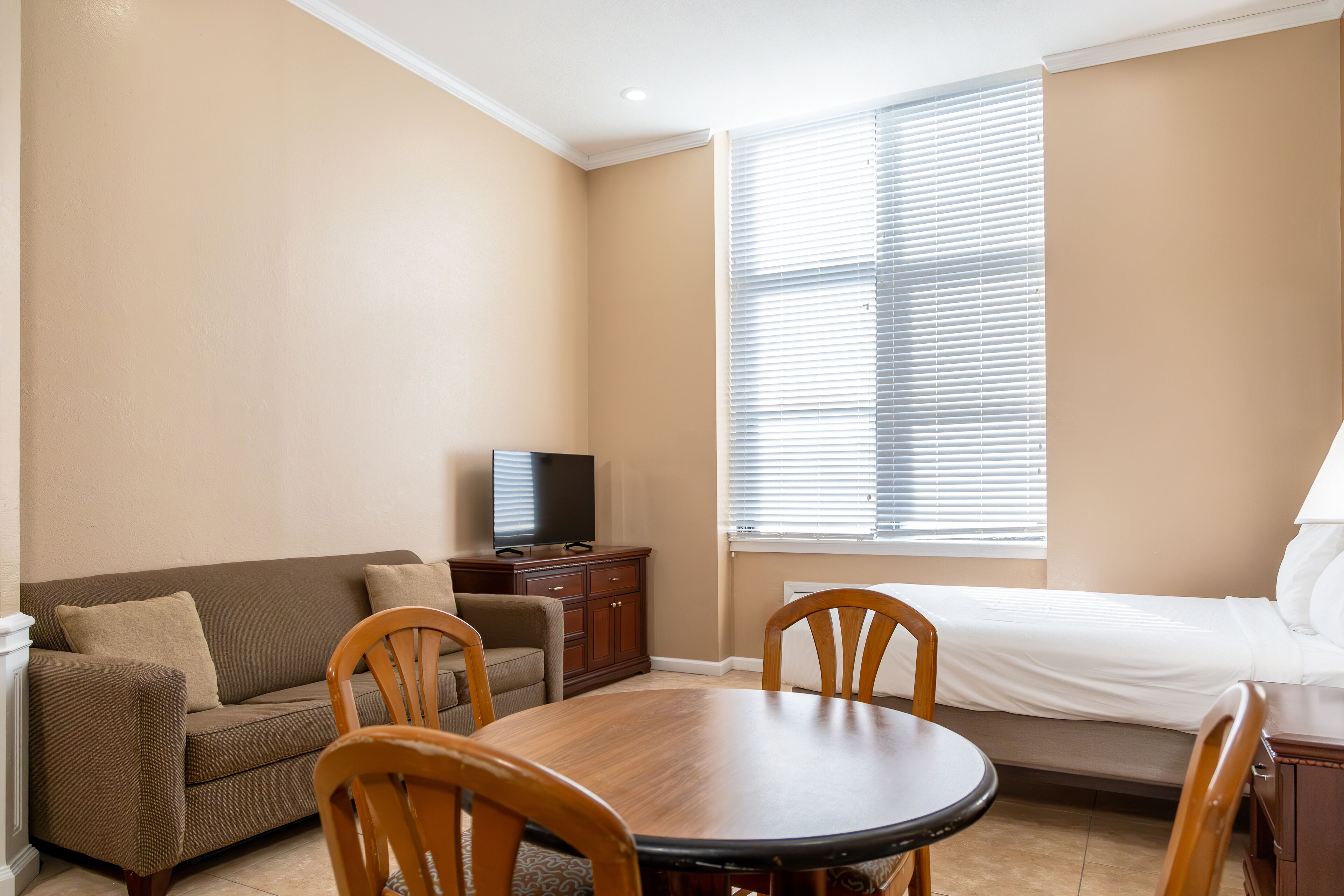 Une chambre d’hôtel avec un canapé, une table à manger avec des chaises, un téléviseur posé sur un meuble et une fenêtre avec des stores.