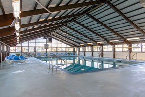 Una piscina interior con paredes de vidrio transparente, sillas reclinables azules y un techo alto con vigas de metal.