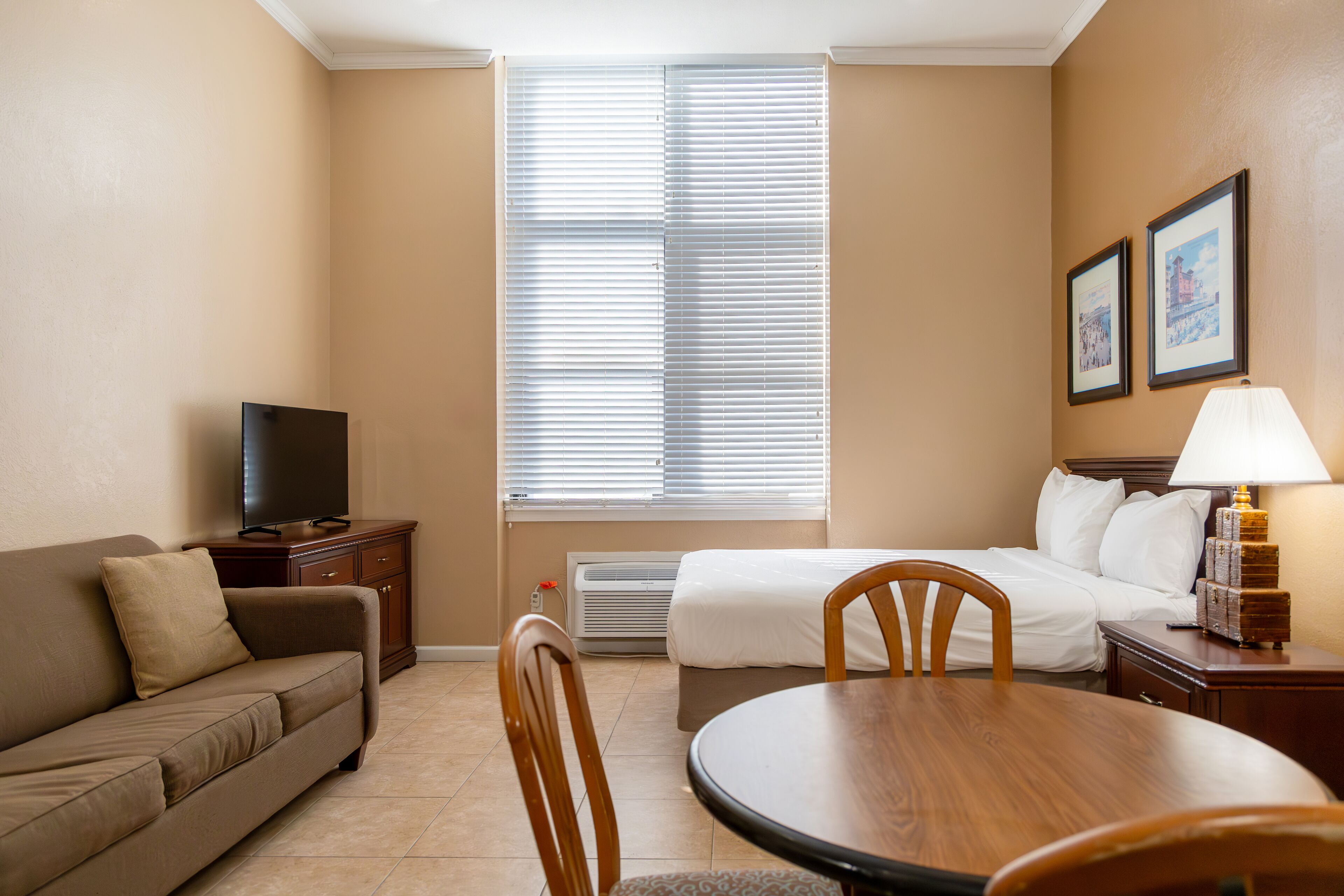 A hotel room with a bed, a TV, a dining table, and a window with blinds.