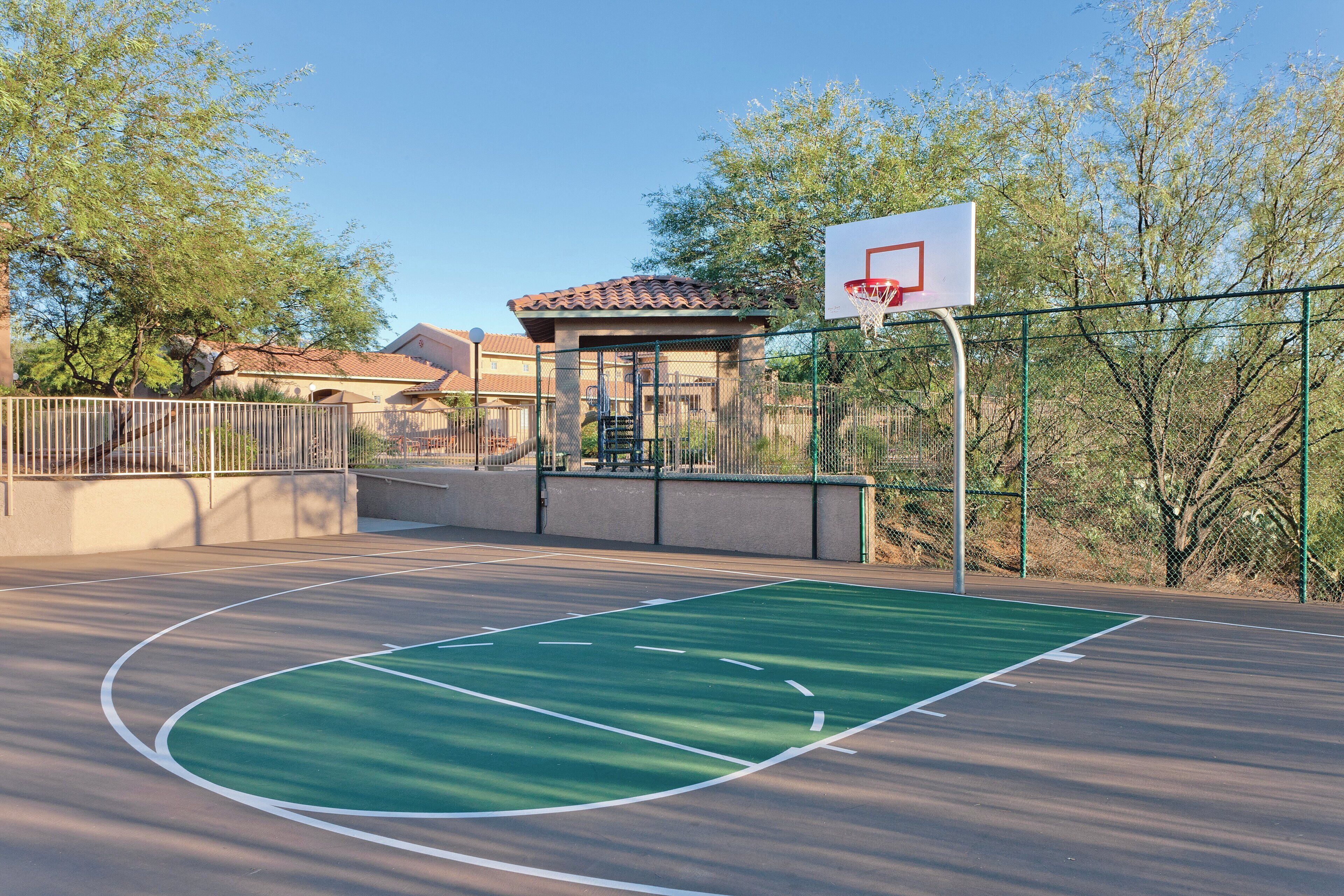 An outdoor basketball court with a green playing surface, a basketball hoop, and a fence.