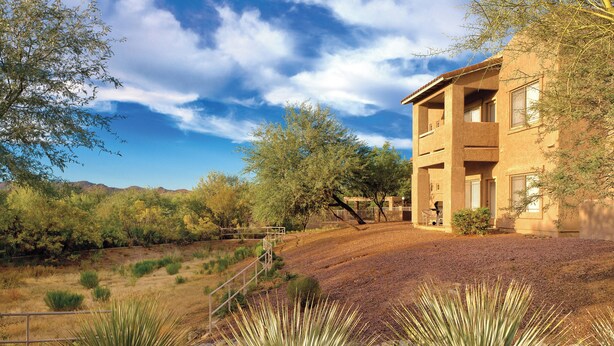 A multi-story building with a flat roof, surrounded by dry grass and yucca plants, set against a backdrop of hills and a clear sky. - WorldMark Rancho Vistoso (Oro Valley)