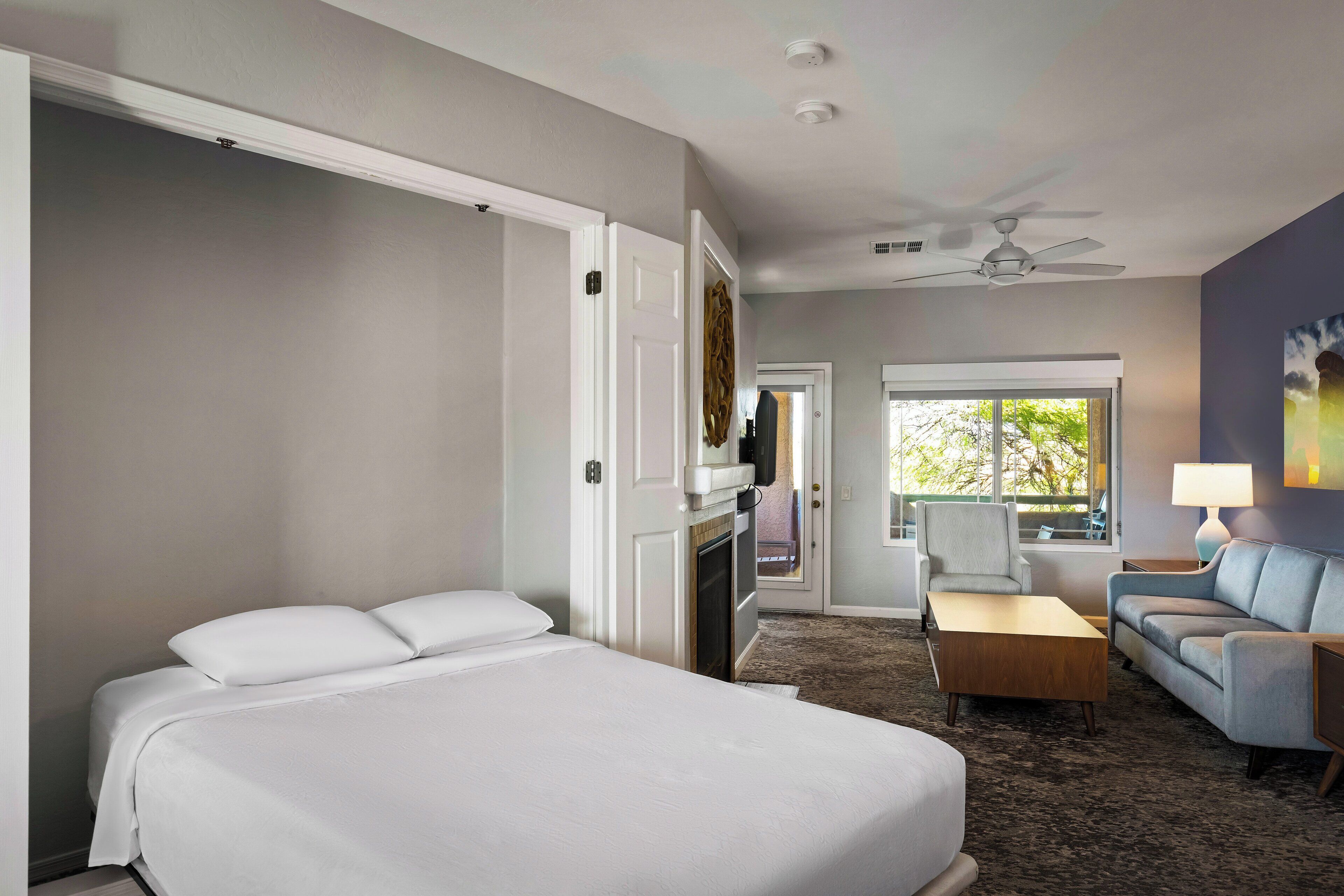 A hotel room with a large bed, a seating area with a sofa and armchair, a small table, and a ceiling fan.