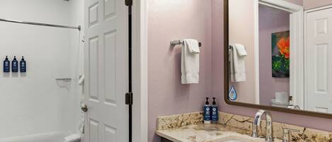 Un baño con una bañera, un lavamanos con encimera de mármol y un espejo grande. Hay dos botellas de champú y una toalla colgada en la pared.