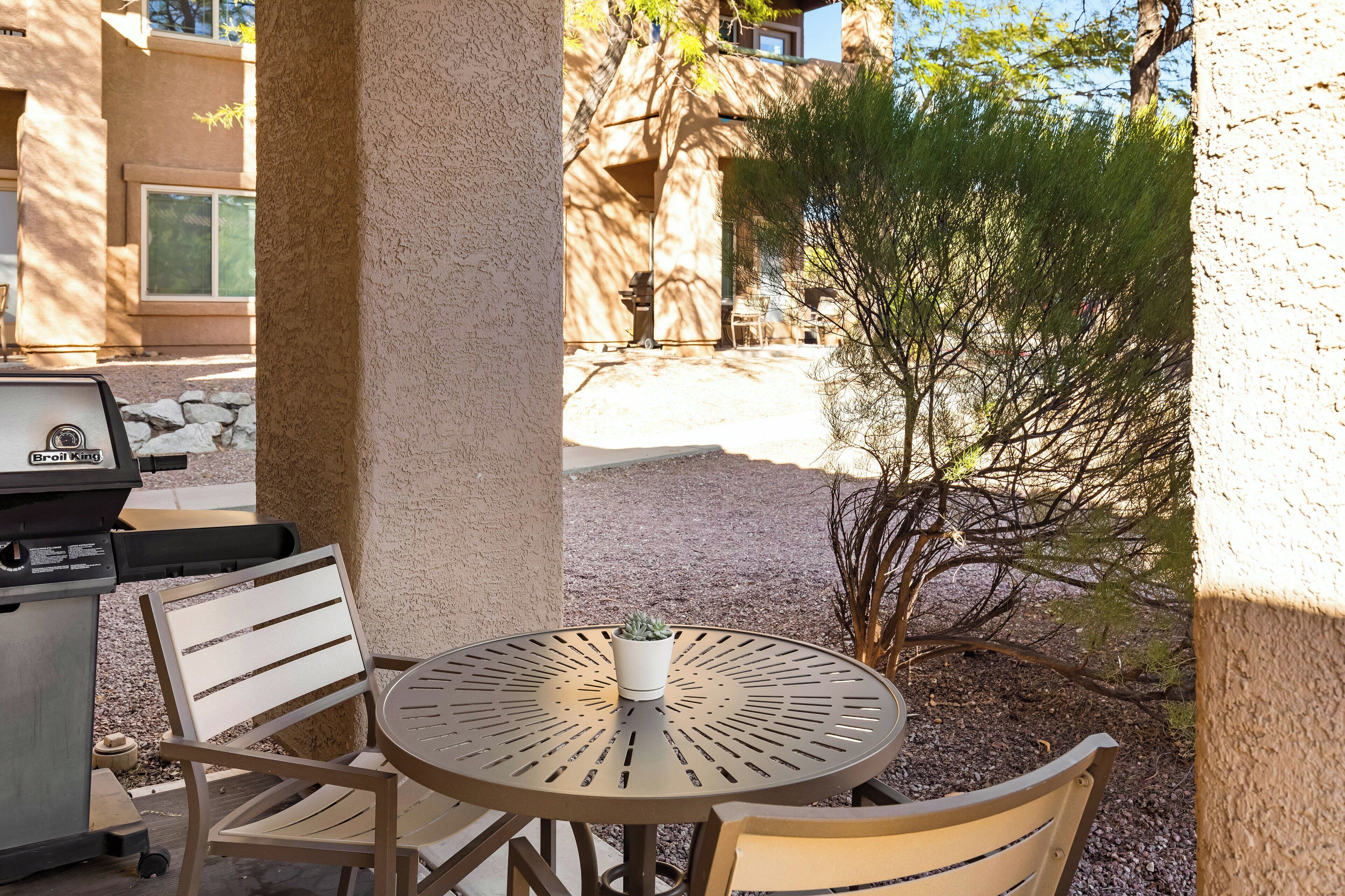 An outdoor patio with a round table, two chairs, a grill, and a small potted plant.