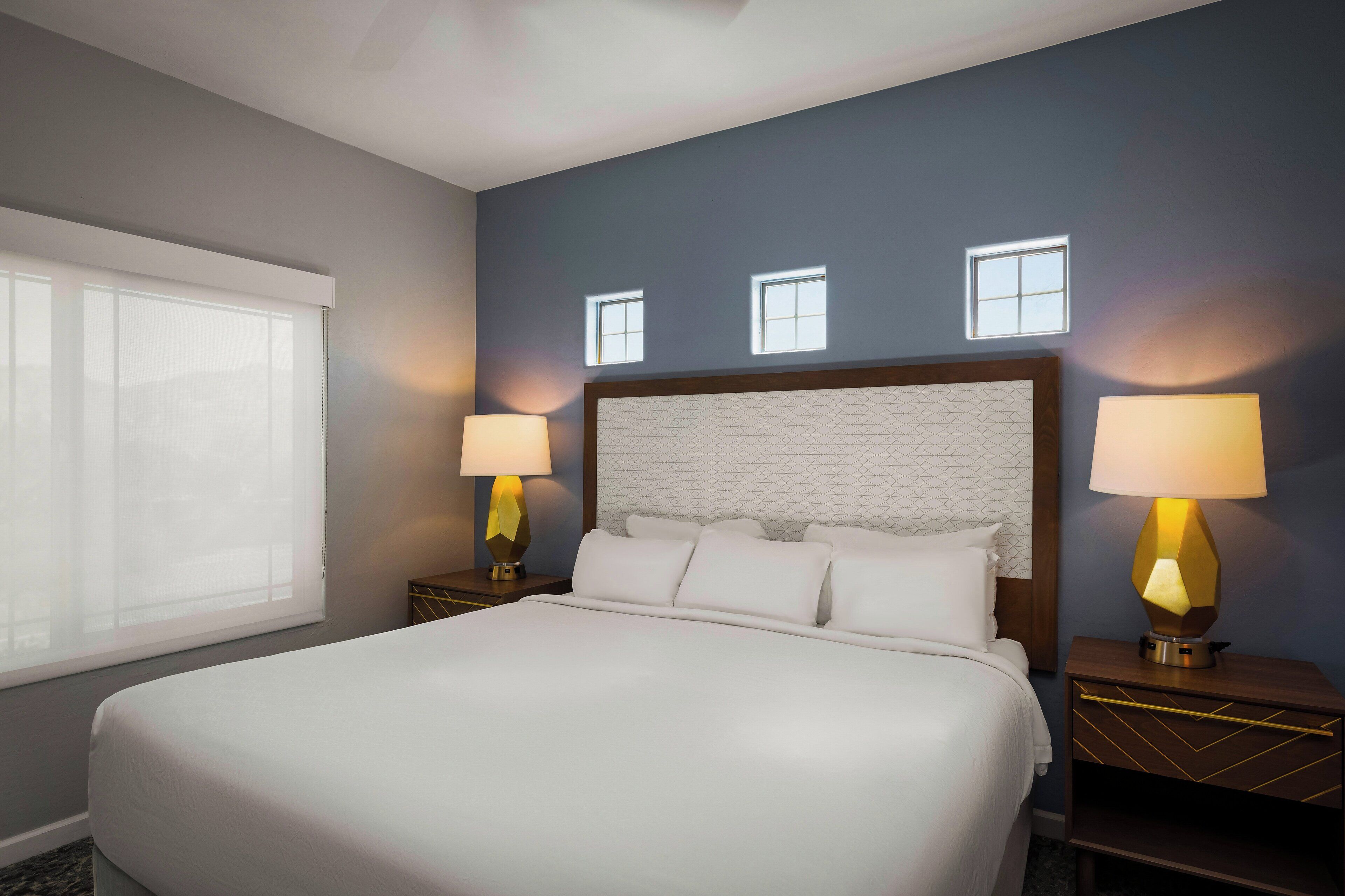 Quarto de hotel com uma cama grande, duas mesas de cabeceira com luminárias, uma janela e uma parede com três pequenas janelas.