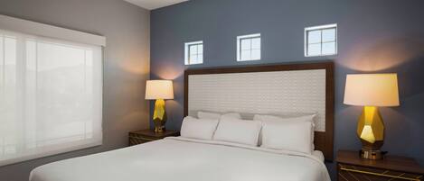 A hotel room with a large bed, two bedside tables with lamps, a window, and a wall with three small windows.