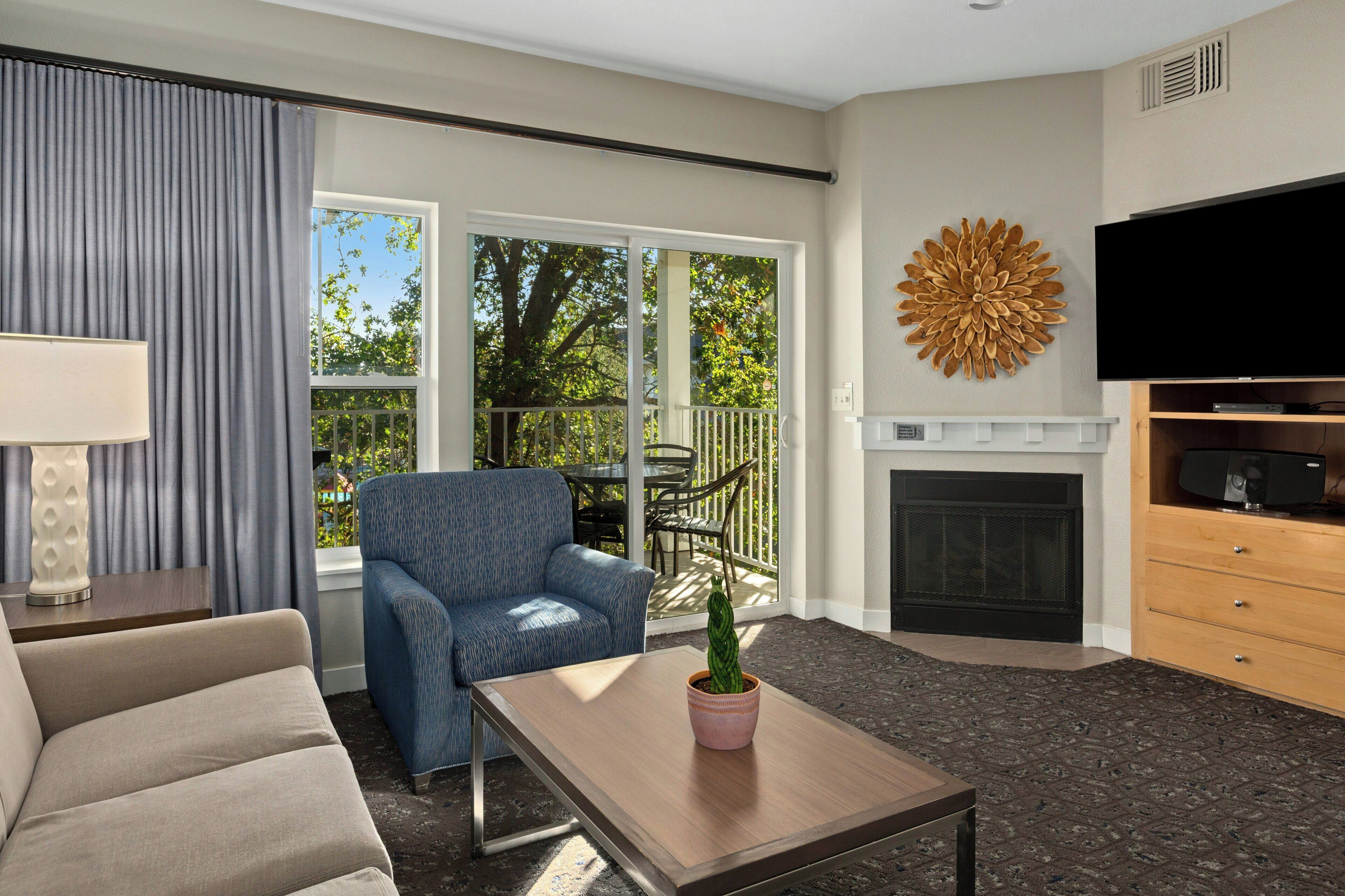 A living room with a sofa, armchair, coffee table, TV, fireplace, and a view of the outdoors.