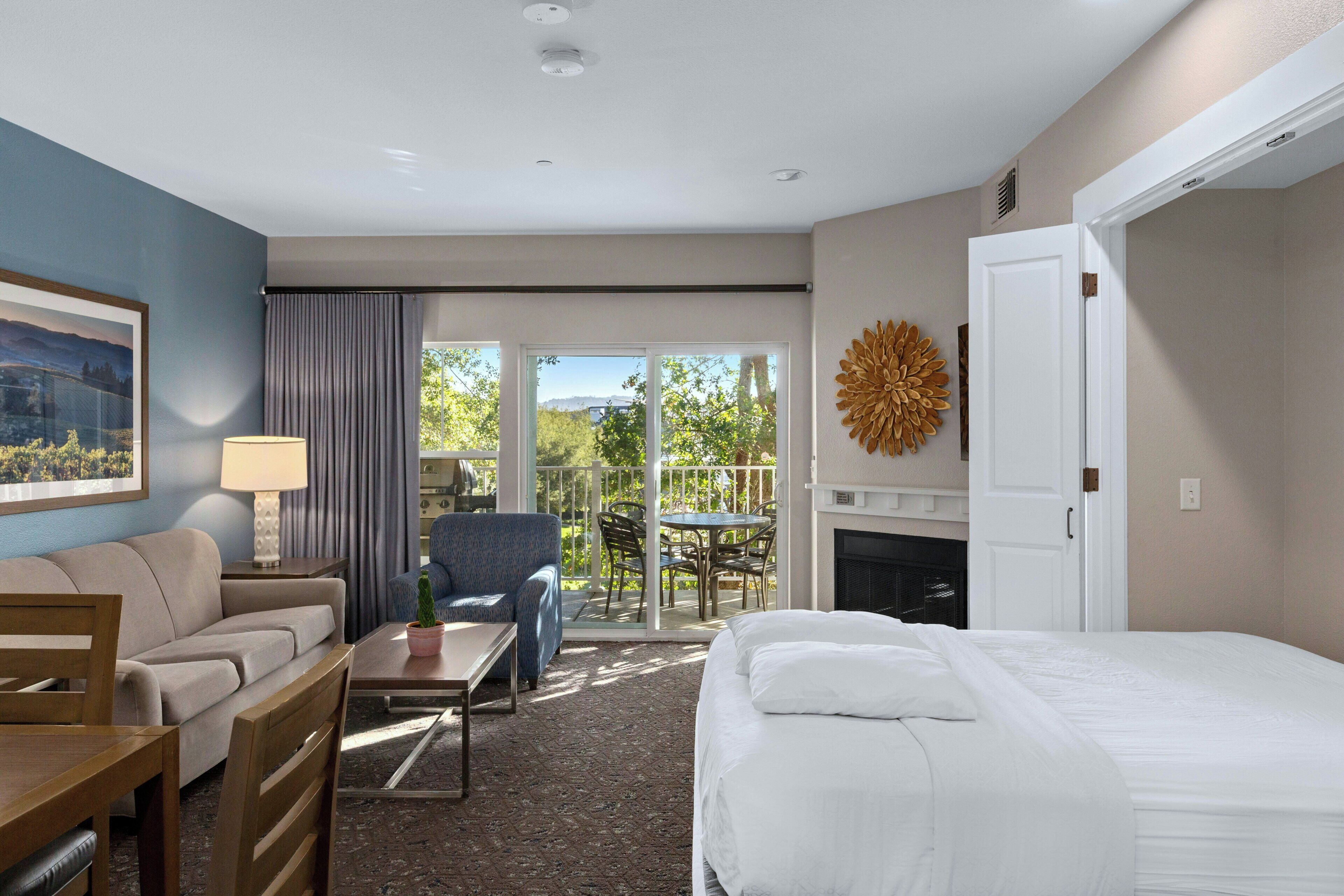 A hotel room with a bed, a sofa, a dining table, a chair, a lamp, a painting, a fireplace, and a view of the outdoors.