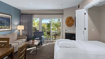 A hotel room with a bed, a sofa, a dining table, a chair, a lamp, a painting, a fireplace, and a view of the outdoors.
