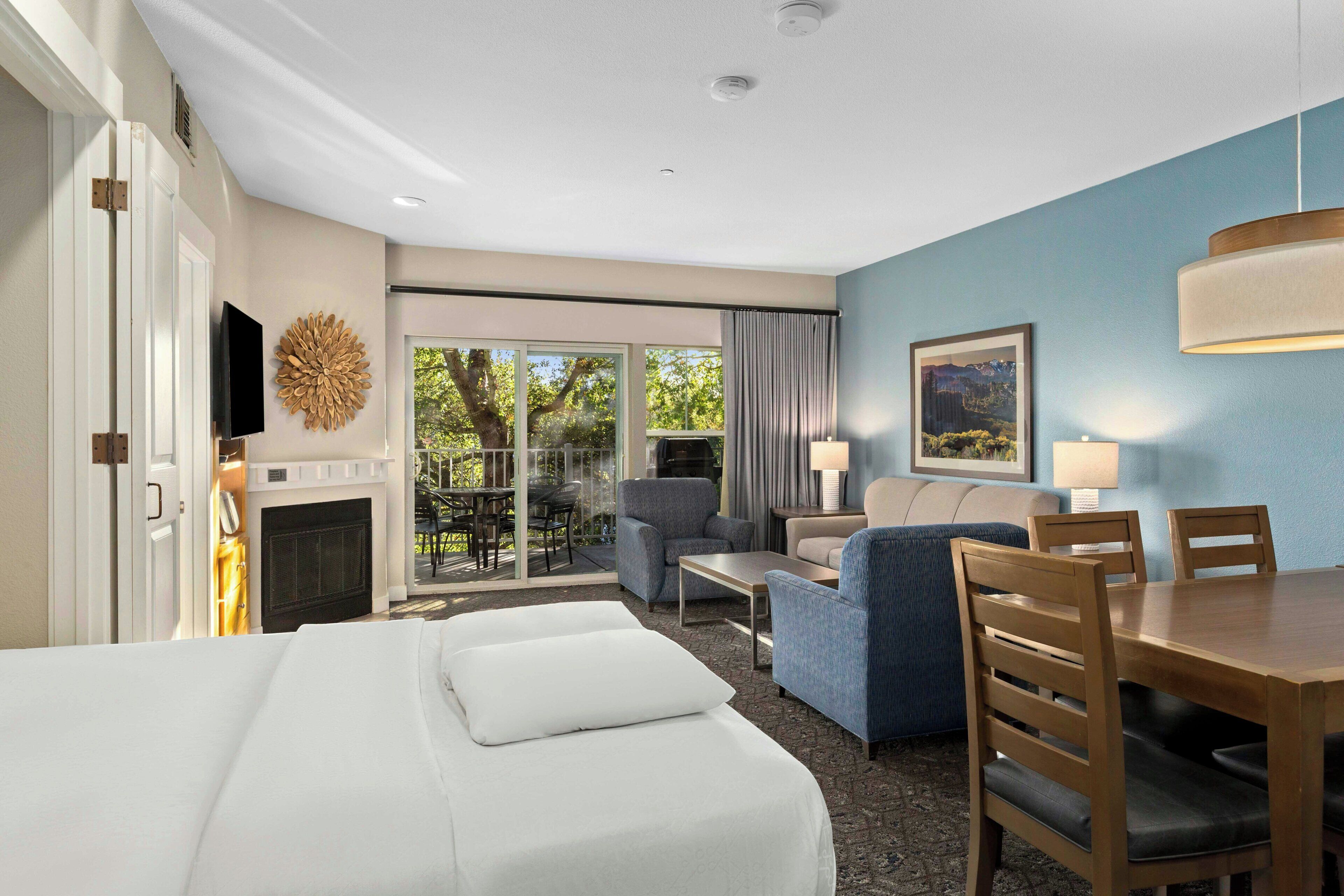 A hotel room with a bed, a seating area with a sofa and chairs, a dining table, a fireplace, and a large window with a view of trees.