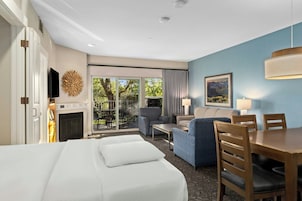 A hotel room with a bed, a seating area with a sofa and chairs, a dining table, a fireplace, and a large window with a view of trees.