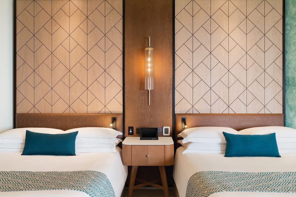 Habitación de hotel con dos camas, una mesita de noche con panel de control y un diseño de panel en la pared.