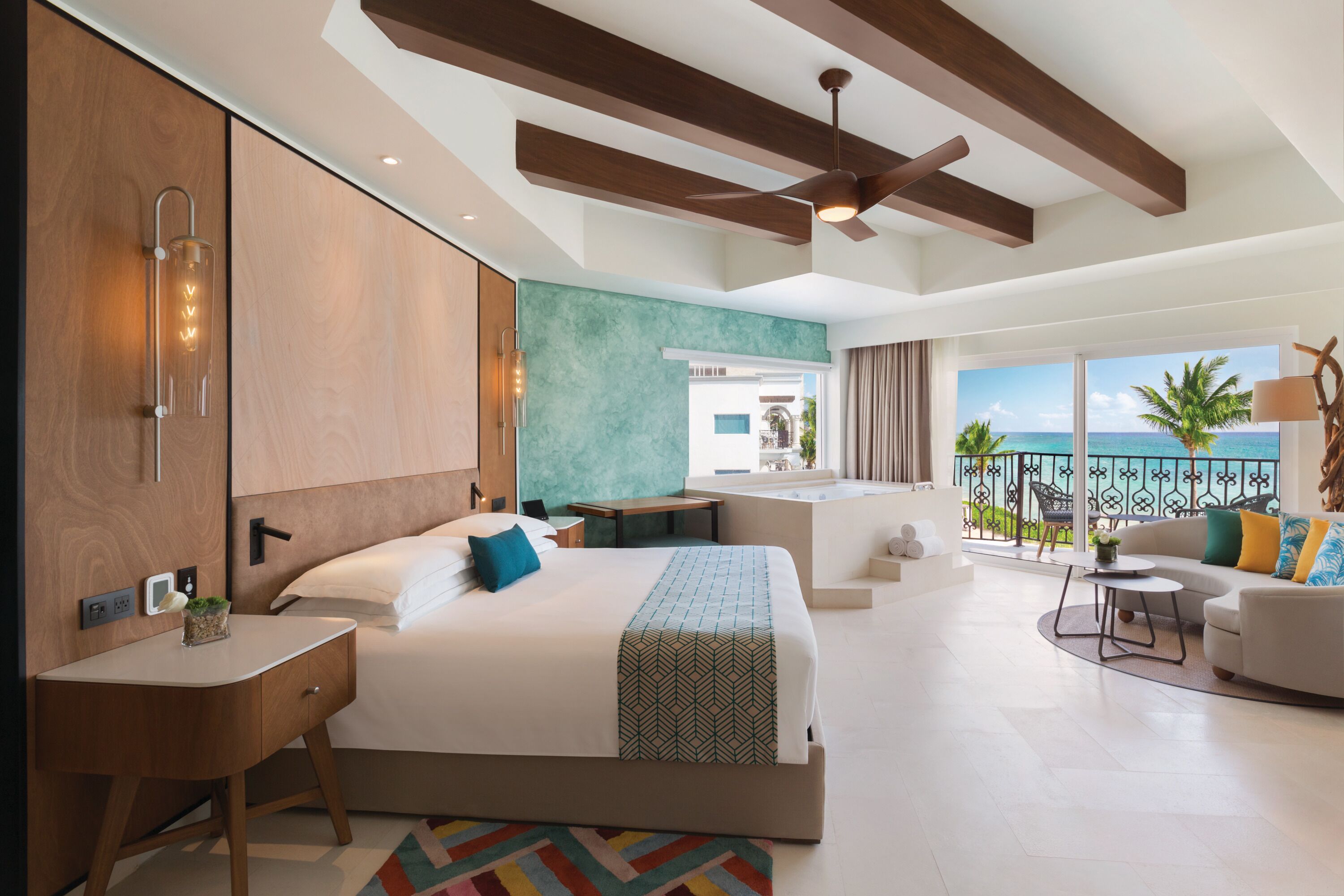 A modern hotel room with a large bed, a desk, a balcony with a view of the ocean, and a ceiling fan.