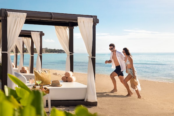 Una cabaña en la playa con una cama, toallas y una bebida sobre la mesa.