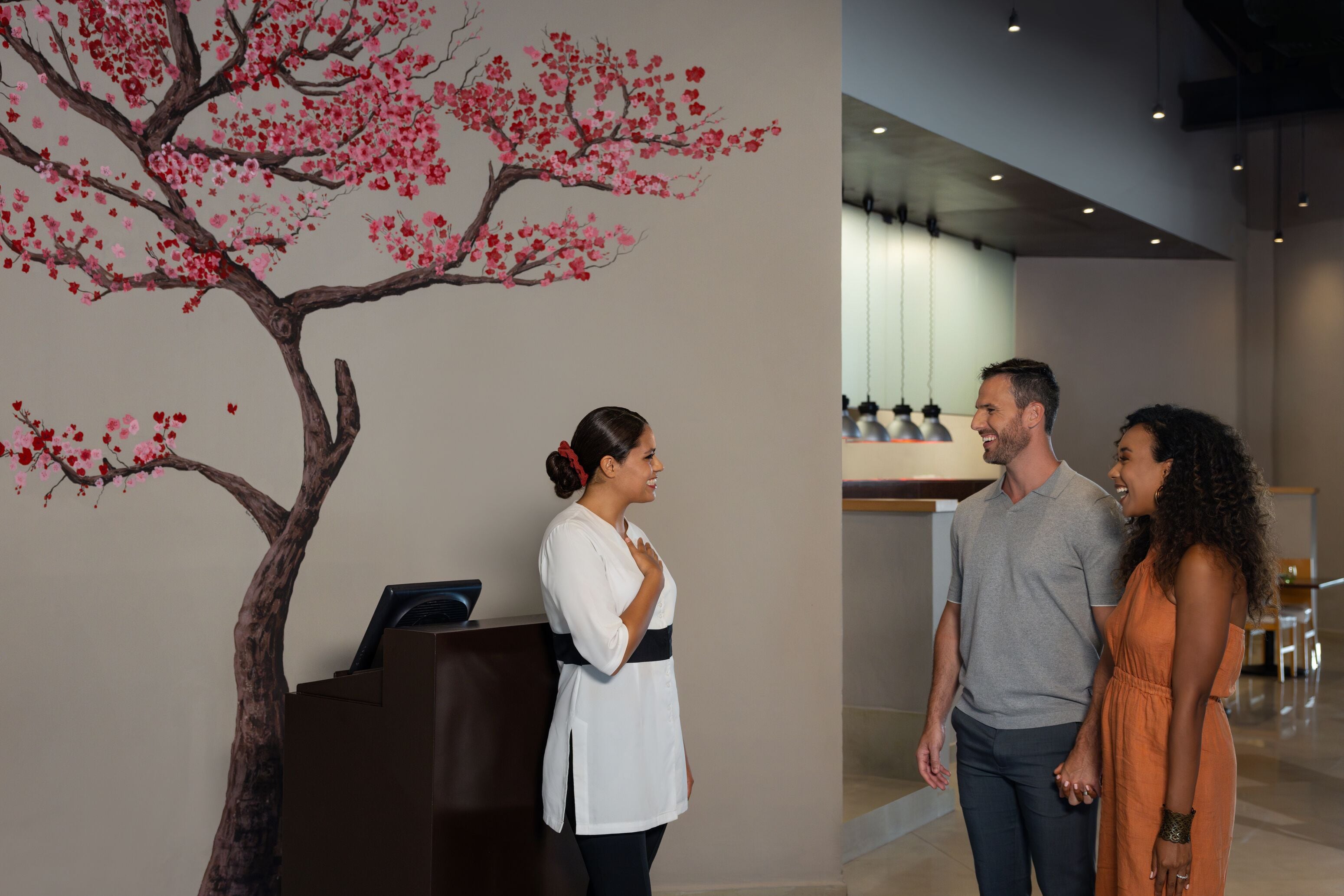 A reception desk with a wall mural of a tree with pink blossoms, a modern interior with a high ceiling, and a seating area with tables and chairs.