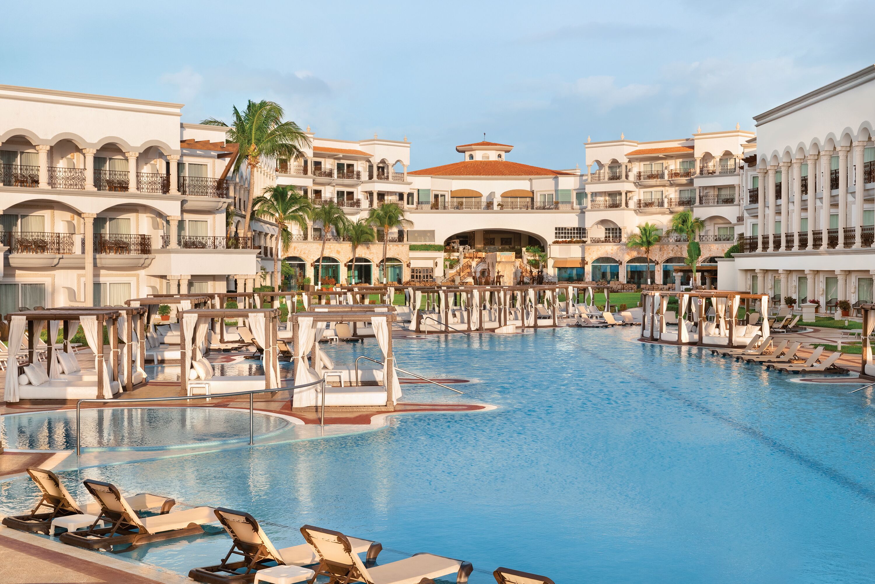Zona de piscina en un resort con múltiples sillas de descanso y sombrillas, rodeada de edificios de varios pisos con balcones y una estructura central.