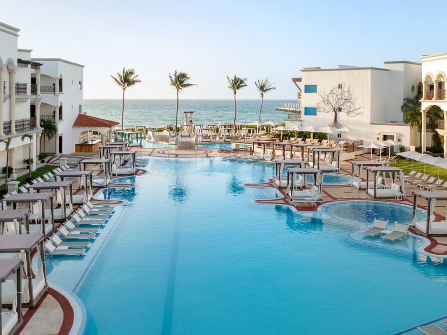Una gran piscina con varias zonas de descanso y palmeras, rodeada de edificios con balcones y vista al océano.
