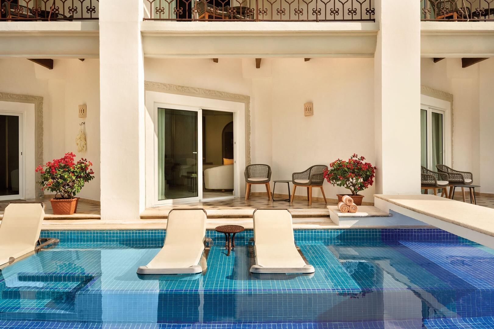 A pool area with lounge chairs, potted plants, and a view of the interior.
