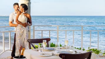 A couple embracing at a table with a view of the sea, a white railing, and a bottle of champagne.