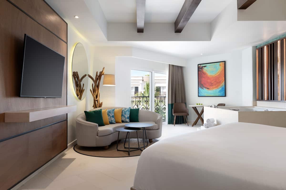 Una habitación de hotel moderna con una cama grande, un sofá con cojines de colores, una mesita redonda pequeña, un televisor montado en la pared y un cuadro en la pared.