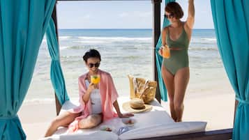 Una cabaña frente a la playa con vista al océano, una tumbona y una mesa de comedor con una bebida, un sombrero de paja y una cesta.