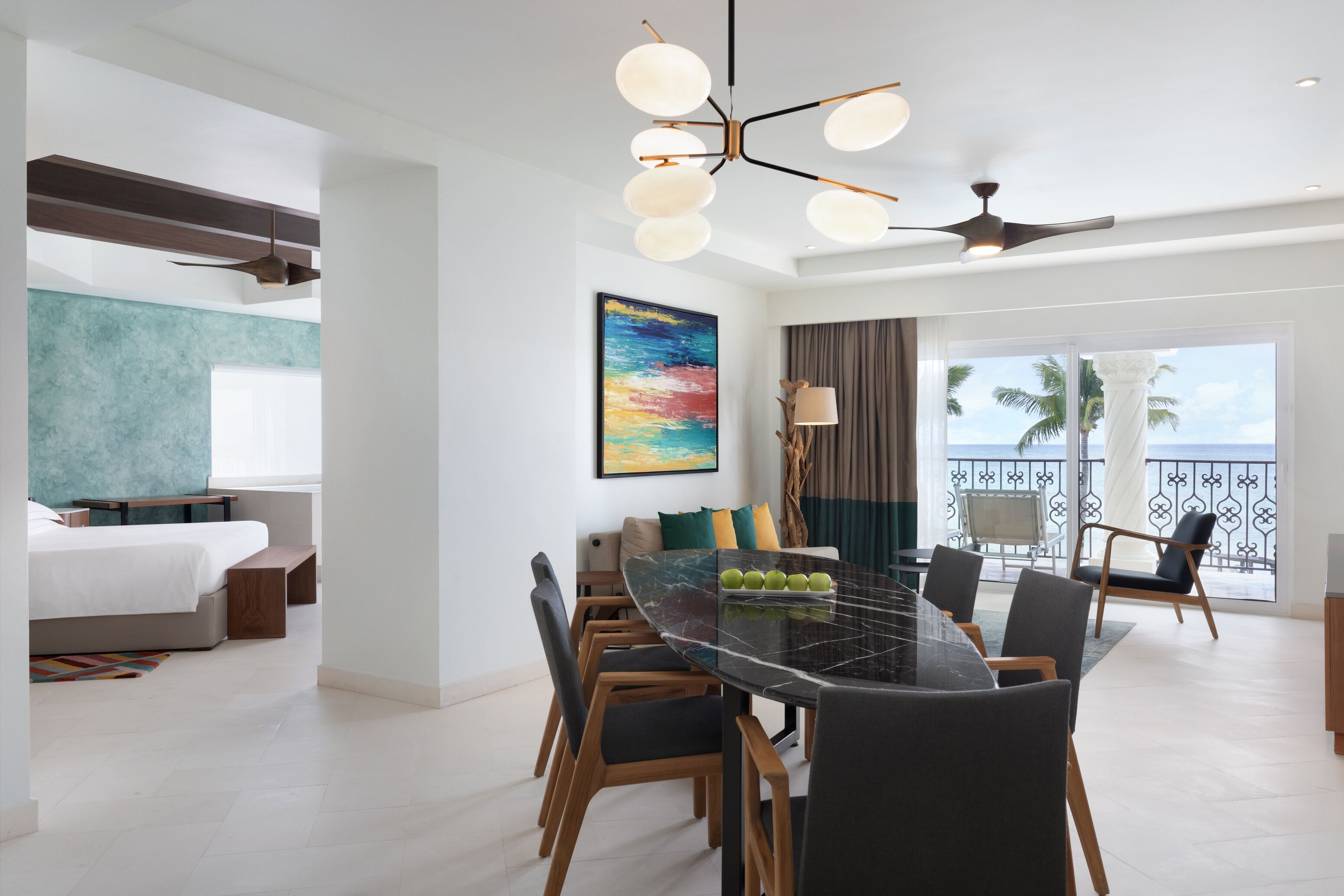 A modern hotel room with a dining area, a large painting, and a balcony with a view.