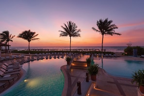 Zona de piscina con sillas de descanso y palmeras, con vista al océano al atardecer.