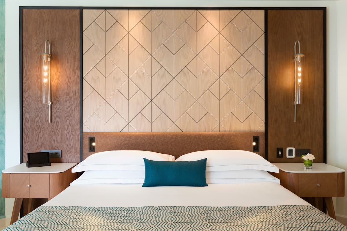 Una habitación de hotel con una cama grande, dos mesitas de noche, un velador con lámpara y un panel de pared con diseños geométricos.
